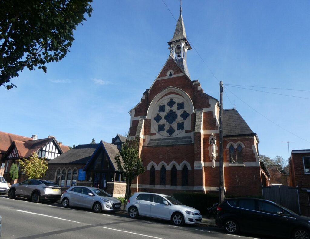 The red-brick Catholic Church of St Mary Immaculate on West Street, Warwick, with a rose window and bell turret, and cars parked along the street outside