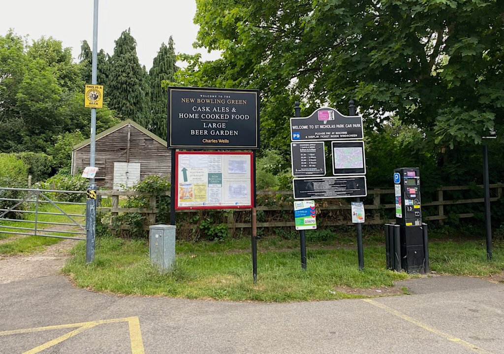 The St Nicholas Park car park sign with its tariff board and a pay and display machine beside the entrance, next to the New Bowling Green pub sign, Warwick